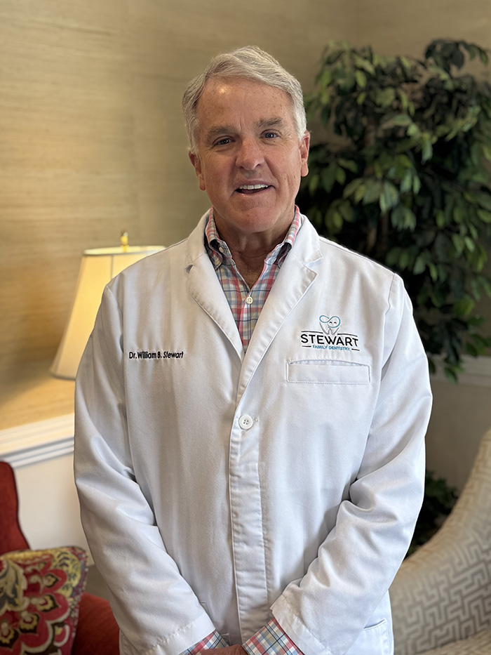 The image shows a man standing indoors, wearing a white lab coat with a name tag that reads  STEWART  and a plaid shirt underneath. He has gray hair and is smiling slightly at the camera.