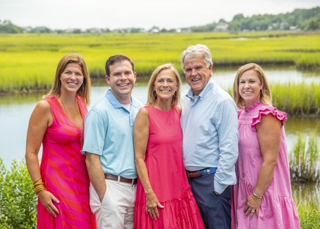 The image shows a group of four adults posing together outdoors near a body of water, with a backdrop of greenery and a cloudy sky.