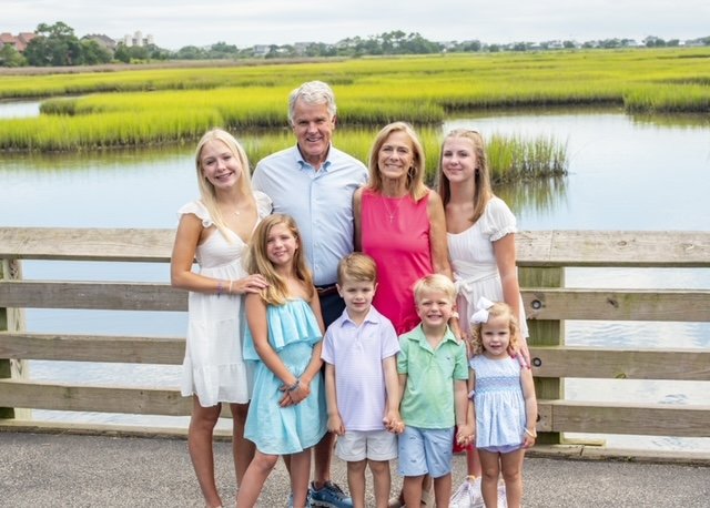 The image shows a family posing outdoors on a wooden deck by a body of water, with a man standing behind them and a woman sitting in front. They are dressed for a warm day, and all members of the family are smiling at the camera.