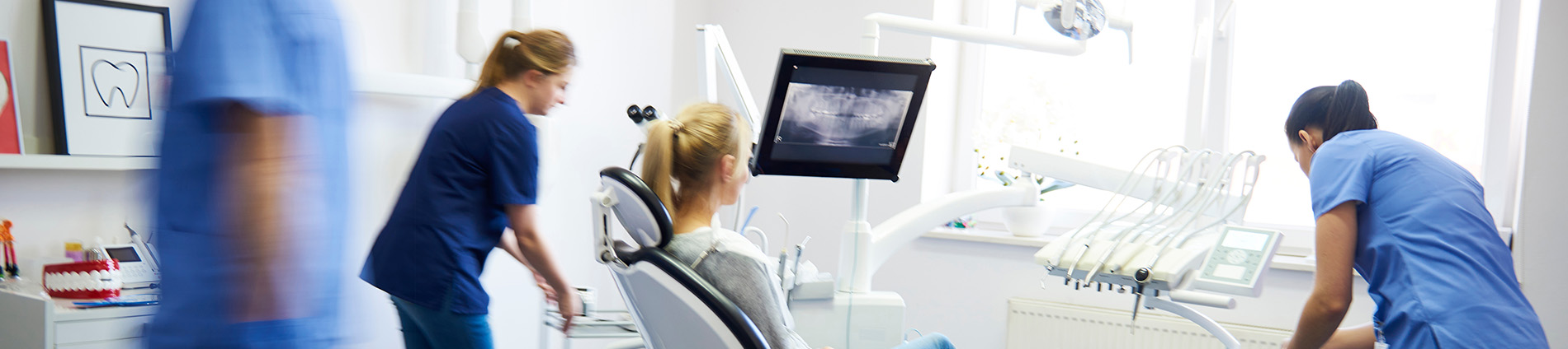 The image shows a dental office setting with two individuals wearing medical scrubs standing near a dental chair, engaged in their work.
