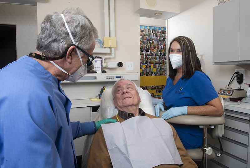 In the image, two individuals are engaged in a dental examination in an office setting, with one person seated in a dental chair receiving care, while another person stands behind them, performing the procedure.