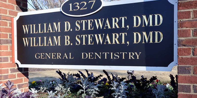 The image shows a sign with the names of three individuals, presumably dentists, in front of a brick building, with text indicating their credentials and affiliation with a dental practice.
