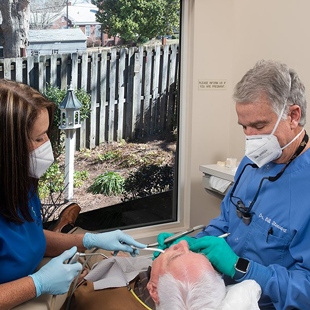 An elderly man receiving dental care from a dentist while a woman in blue scrubs assists, both wearing masks, with a window view of a residential area outside.