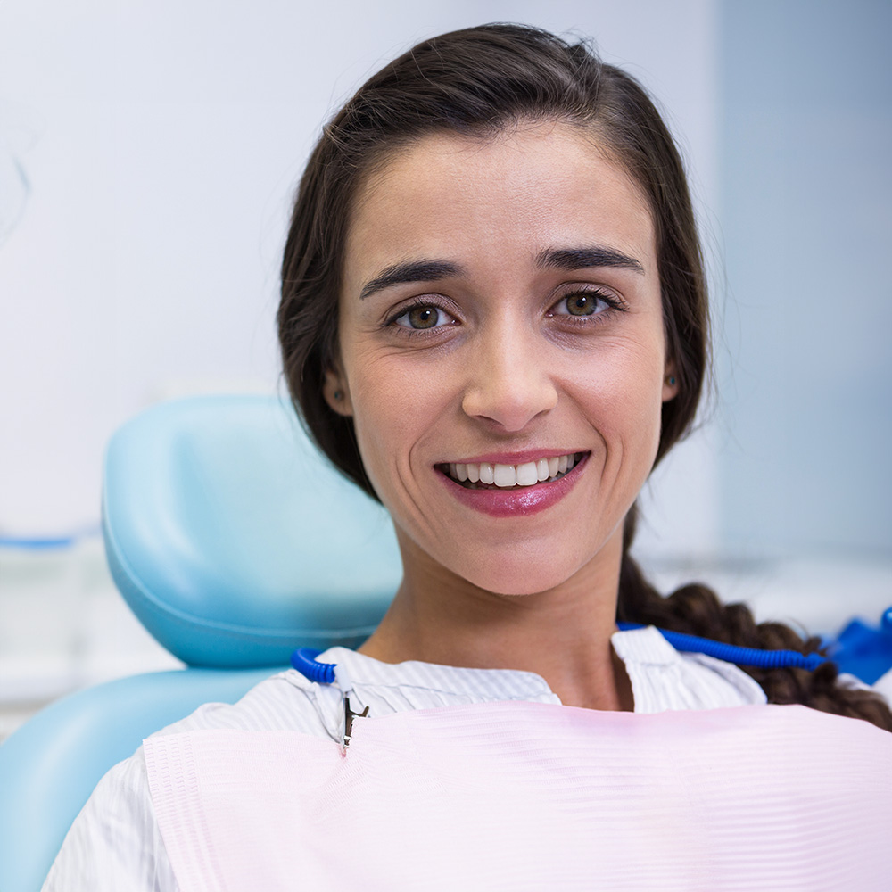 A woman with a broad smile sits in a dental chair, wearing a protective face mask and a blue surgical gown, indicating she is prepared for dental treatment.