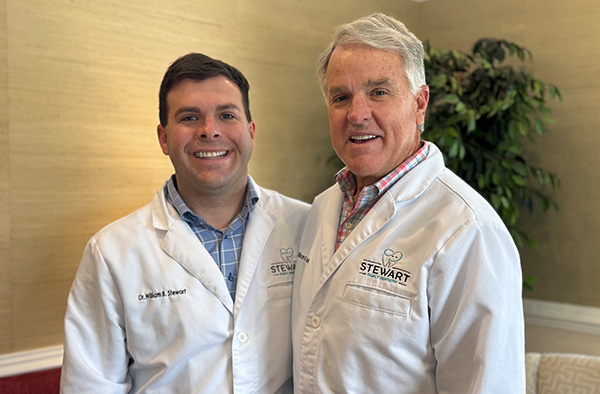 The image shows two men standing side by side  the man on the left is wearing a white lab coat with name tags, while the man on the right is dressed in casual attire. They are posing for a photo indoors, possibly in an office setting.