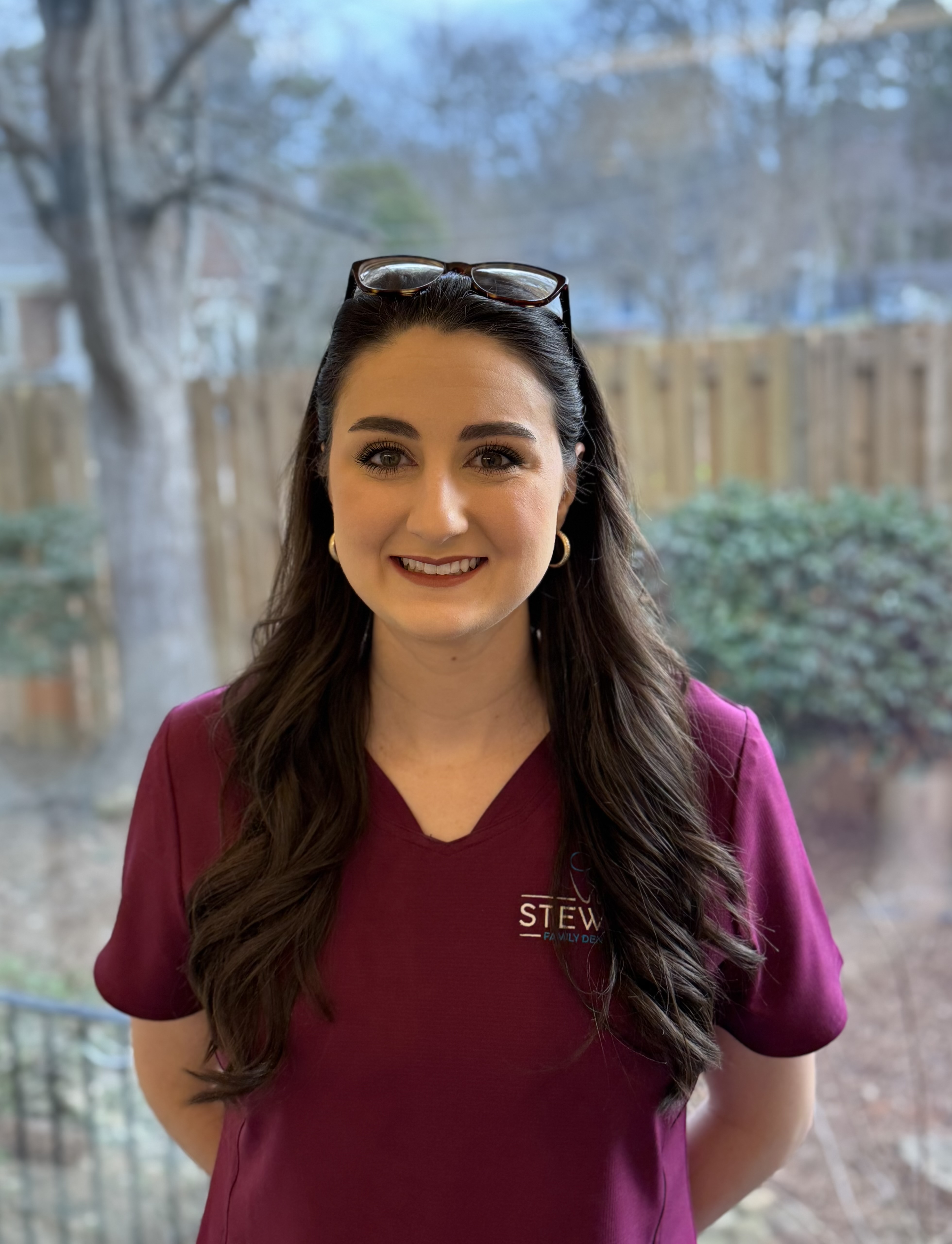 A woman wearing a maroon scrub top stands indoors with a smile, posing for the photograph.