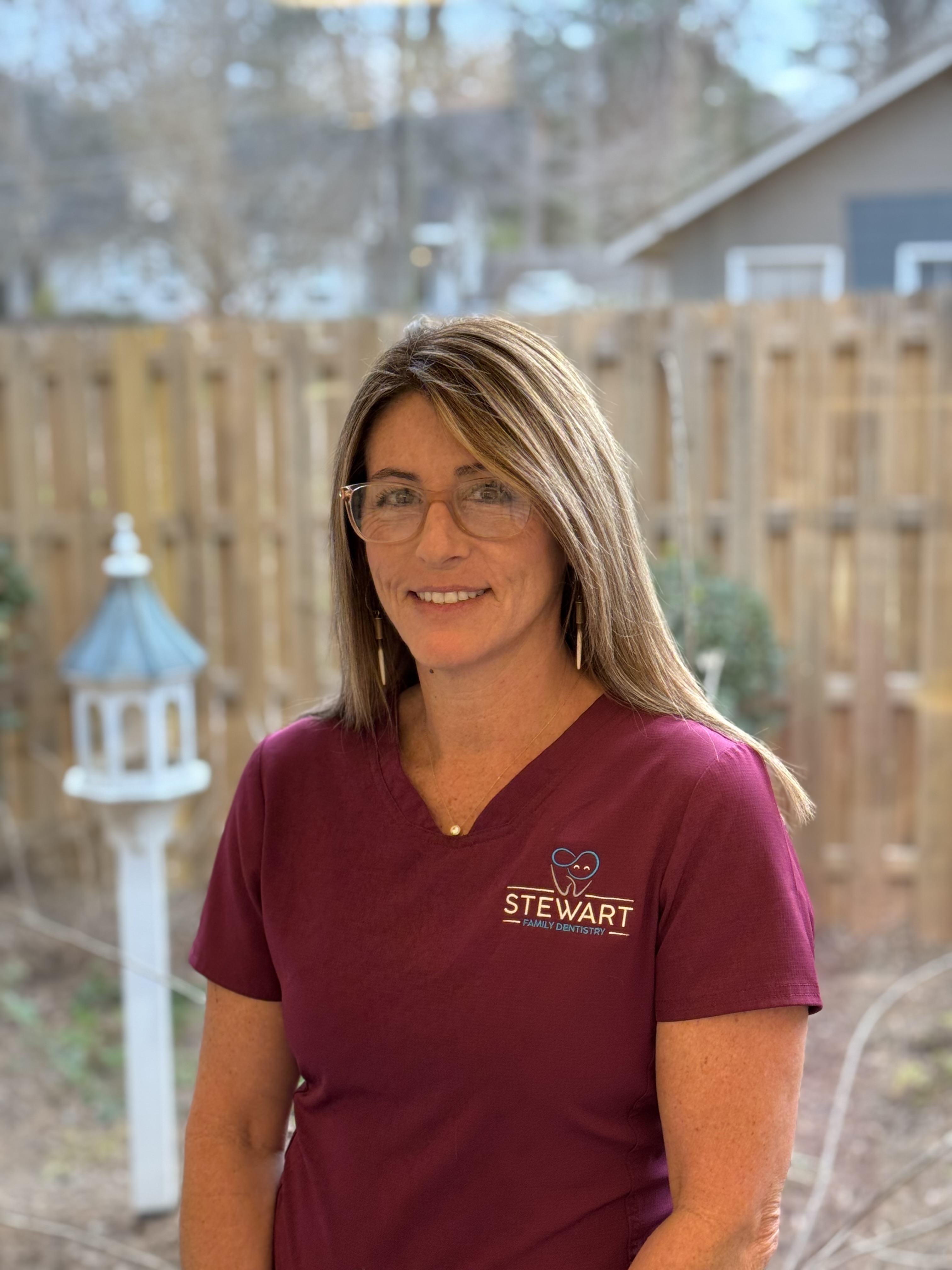 The image shows a woman wearing glasses, a maroon shirt with white text, and a name tag, standing outdoors against a fence and a tree.