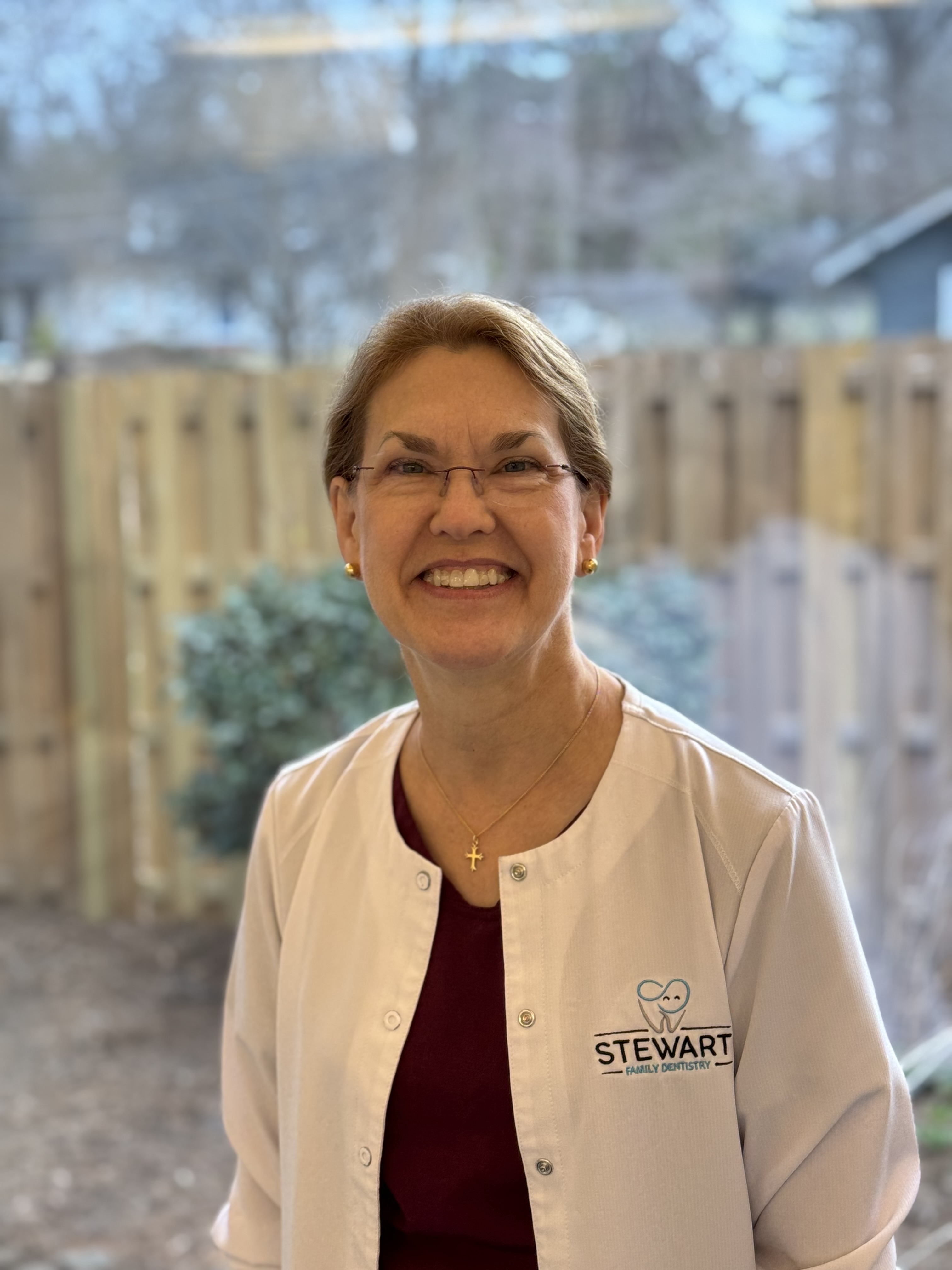 The image shows a woman standing indoors, smiling at the camera, wearing a white lab coat with a logo on the left chest area, and posing for a photograph. She has short hair and appears to be in a professional setting.