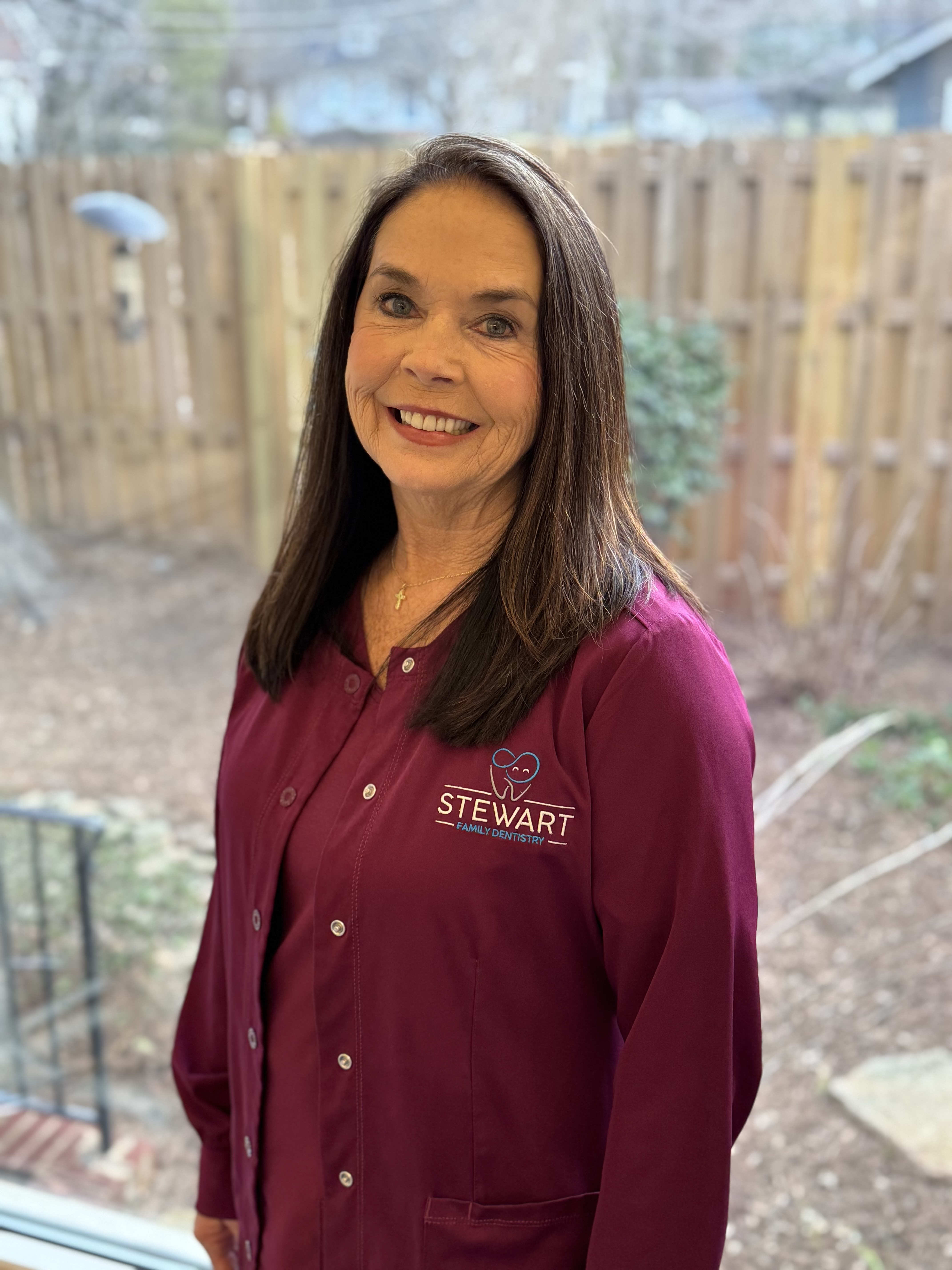 The image shows a woman standing indoors, wearing a blue lab coat with a name tag, smiling at the camera.