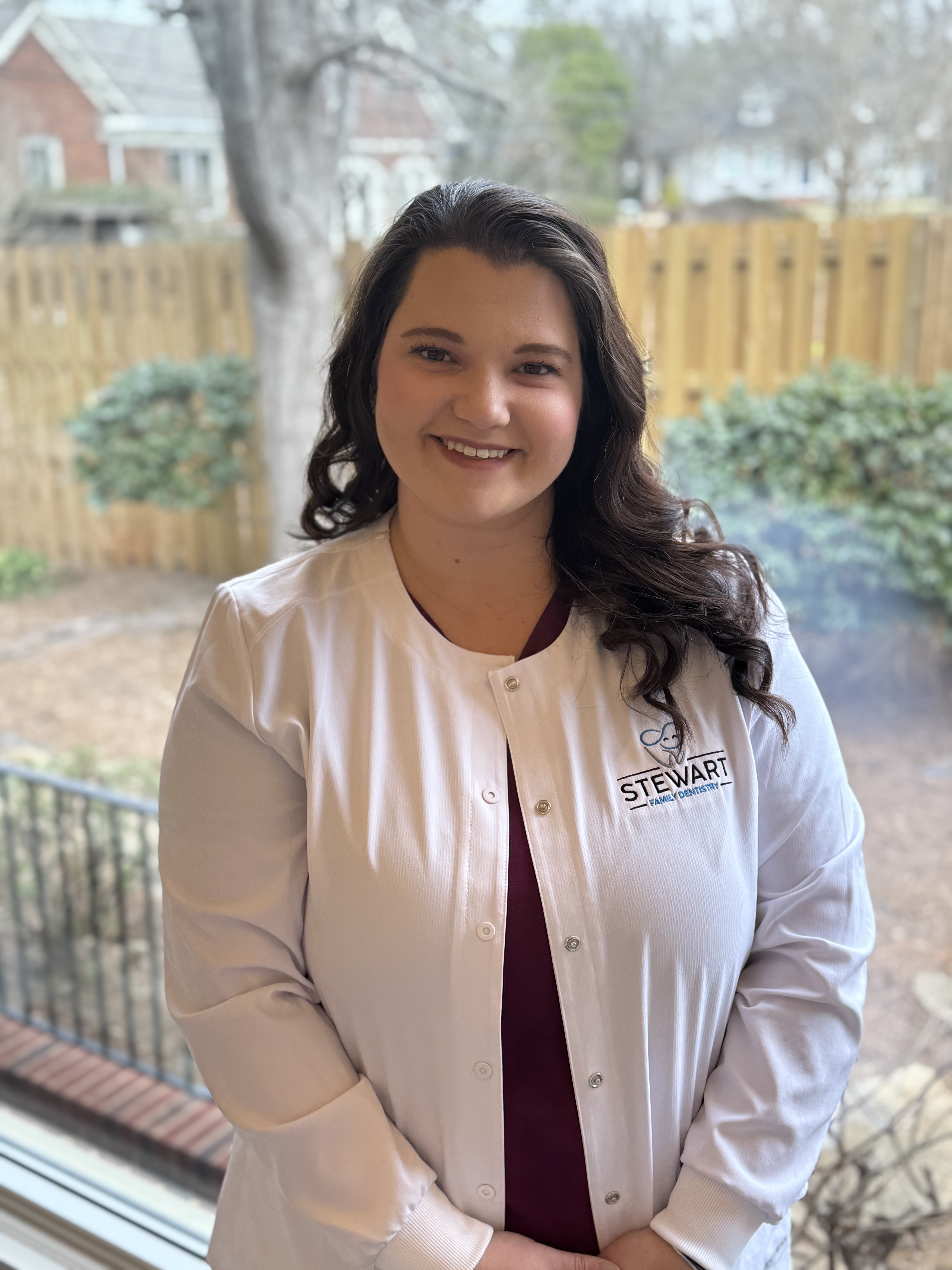 A woman wearing a white lab coat stands in front of a window with a tree outside, smiling at the camera.