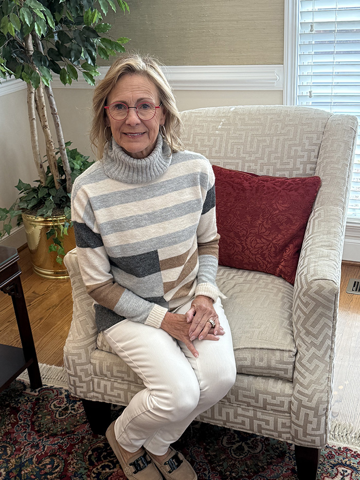 An elderly woman sitting on a chair in an indoor setting, wearing a striped sweater and glasses, with a potted plant visible behind her.