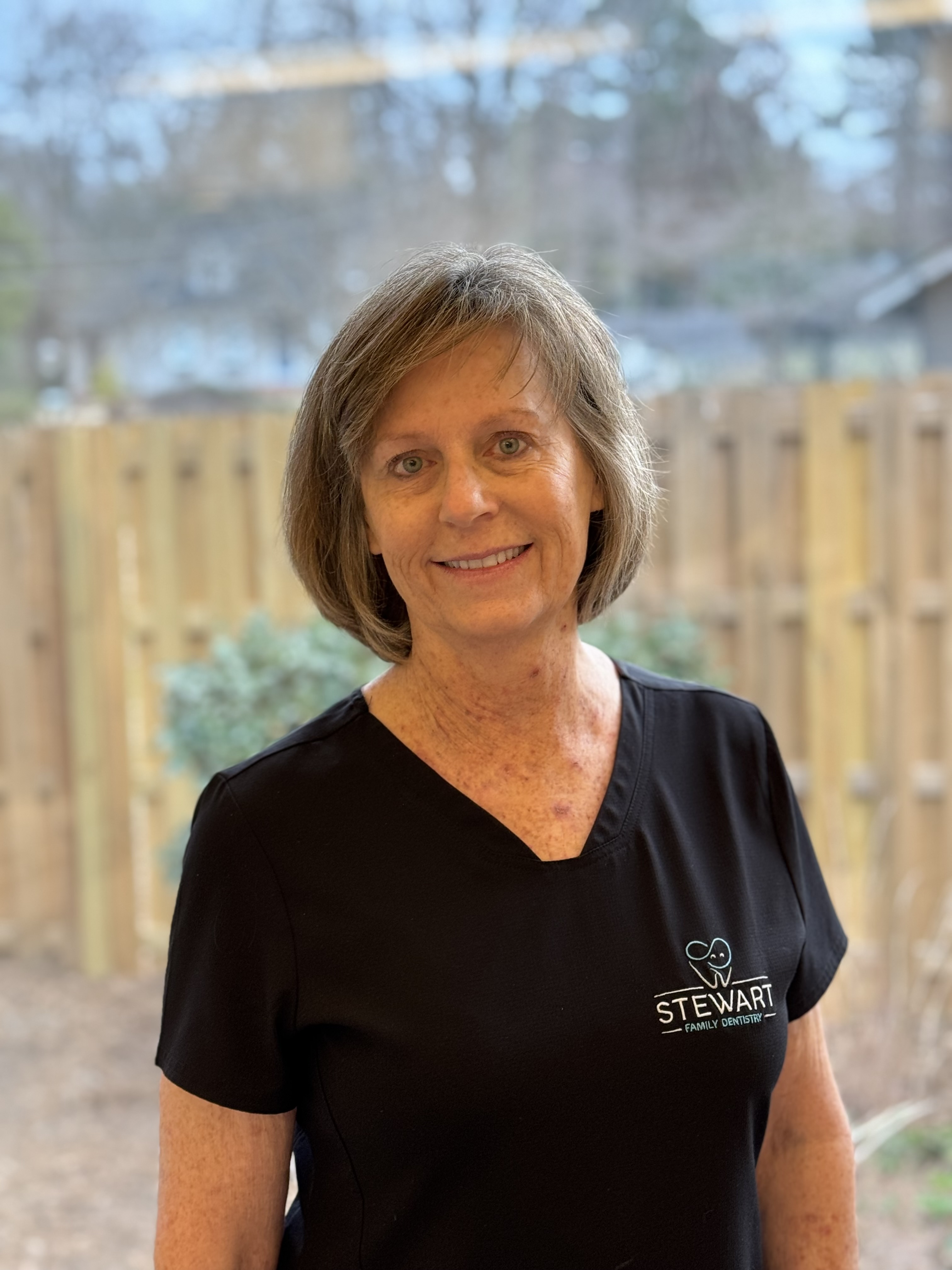 A woman stands in front of a window with blinds, smiling slightly at the camera, wearing a black polo shirt and a lanyard around her neck.
