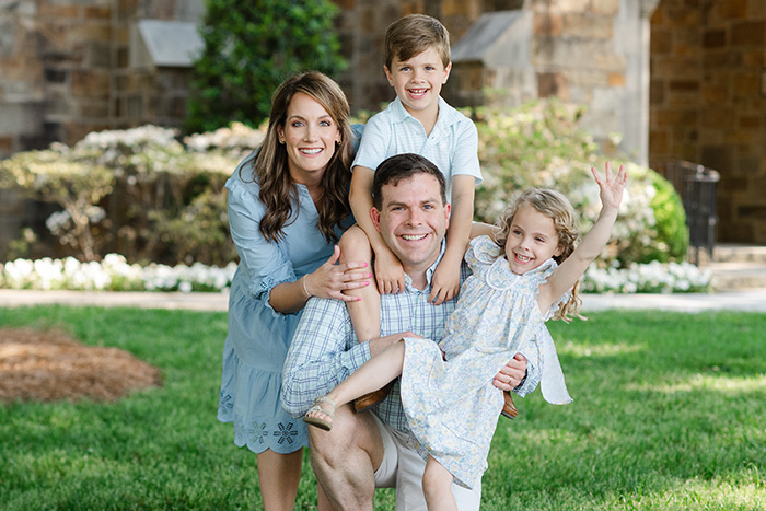 The image shows a family posing outdoors during the daytime  there are four individuals  an adult man, two young children, and one older child. They are dressed casually and are standing on grass in front of a building with a classical architectural style.